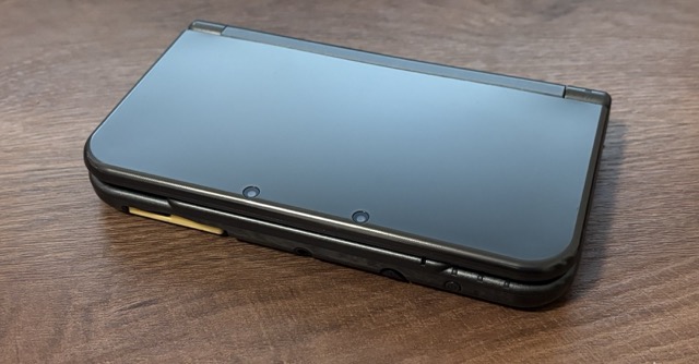 A dark gray-ish Nintendo New 3DS XL, laying on the table, closed.