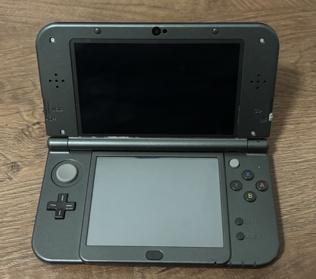 The same Nintendo New 3DS XL as before, laying on the table, opened. The usually dark gray-ish border of this dark gray-ish New 3DS XL looks partly scratched and reveals the gray casing of the LCD.