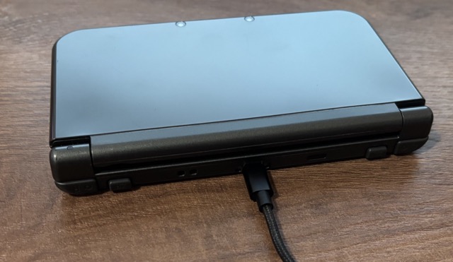 The same Nintendo New 3DS XL as before, laying on the table, closed, turned to its back side.