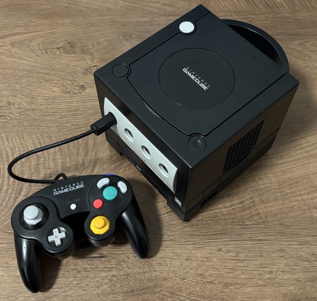 A black Nintendo GameCube, sitting on the table, with a black controller connected to it that's sitting next to it, with its cable curled up underneath the controller.