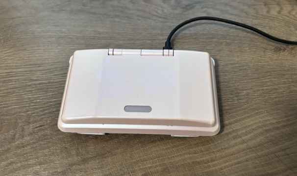 A pink Nintendo DS, laying on the table, closed. A USB-C cable is plugged into the console on its right side from the view of the camera.