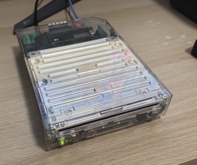 A Nintendo Wii in a clear case, sitting on a desk, with the leg of the monitor it is connected to in frame, turned on. You can very easily see the shielding, airflow covering and even the WiFi antenna for the Retro GEM.